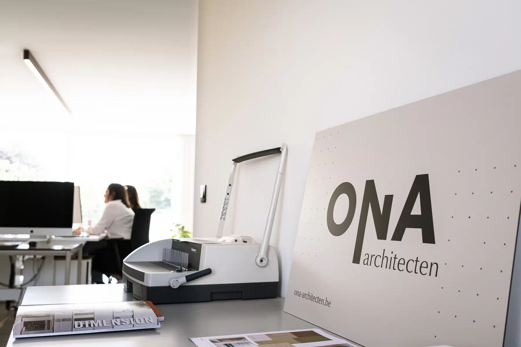 Kantoor van ONA Architecten in Beveren met medewerkster aan het werk achter iMac, en het ONA architecten logo