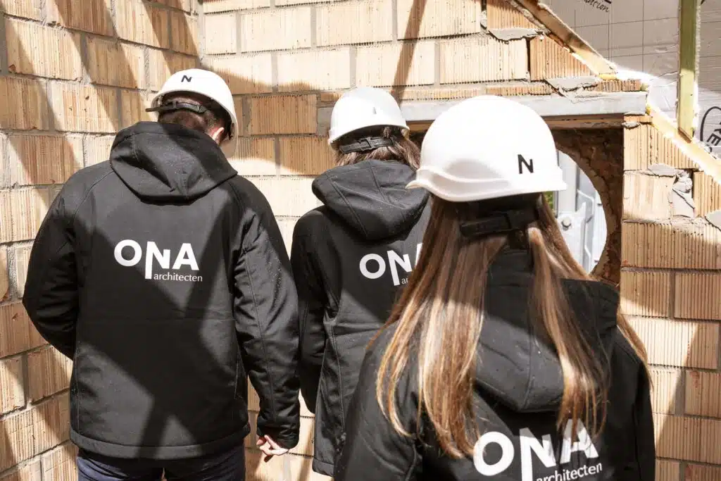 Drie architecten van ONA Architecten in zwarte jackets en witte helmen inspecteren een bouwwerf in Beveren.