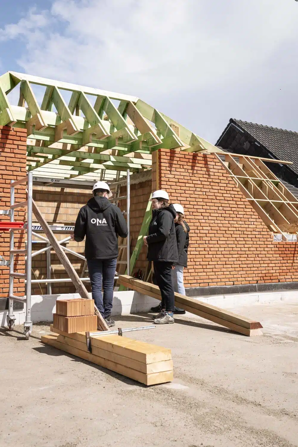 Drie architecten van ONA Architecten in zwarte jas en witte helm bespreken de werfopvolging bij een bakstenen gebouw