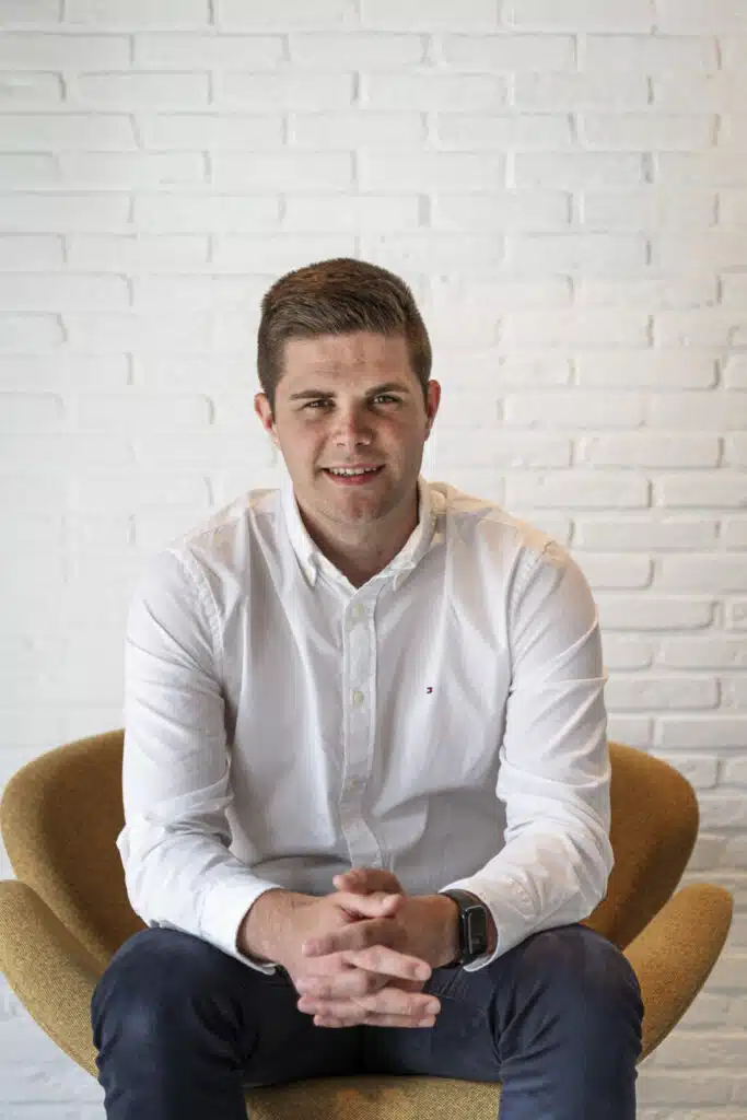 Michiel, architect bij ONA Architecten Beveren, zit glimlachend in een gele stoel voor een witte bakstenen muur