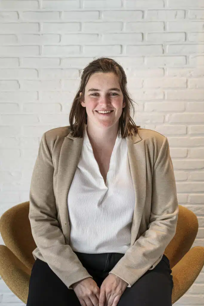 Flore, architect bij ONA Architecten in Beveren, zittend voor een witte bakstenen muur in een beige blazer en witte blouse