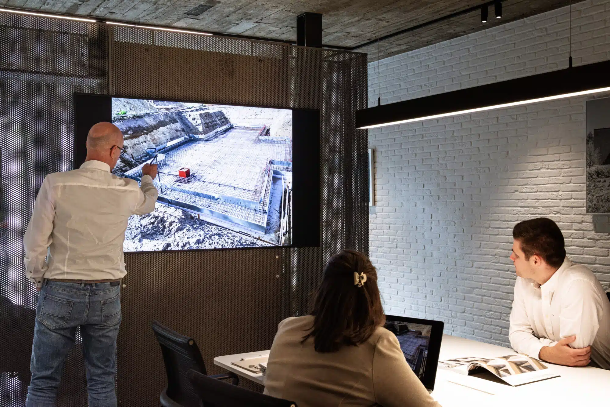 Architect van ONA Architecten presenteert bouwplaatsfoto op groot scherm tijdens projectbespreking met collega's in modern ka